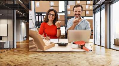 Middle age couple working at small business ecommerce pointing to the back behind with hand and thumbs up, smiling confident Wall mural