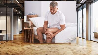 Middle age grey-haired man suffering for knee injury sitting on bed at bedroom Wall mural