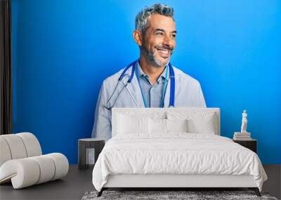 Middle age grey-haired man wearing doctor uniform and stethoscope looking away to side with smile on face, natural expression. laughing confident. Wall mural