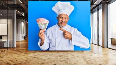 Middle age grey-haired man wearing professional cook uniform holding new zealand dollars banknotes smiling happy pointing with hand and finger Wall mural