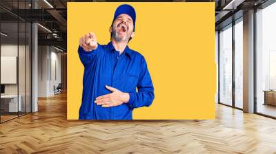 Middle age handsome man wearing mechanic uniform laughing at you, pointing finger to the camera with hand over body, shame expression Wall mural