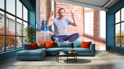 Middle age handsome sportman sitting on mat doing stretching yoga exercise at gym smiling amazed and surprised and pointing up with fingers and raised arms. Wall mural