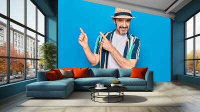 Middle age handsome tourist man on vacation wearing shirt and hat over blue background smiling and looking at the camera pointing with two hands and fingers to the side. Wall mural