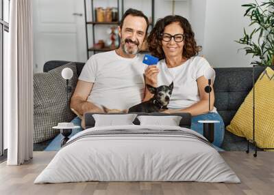 Middle age hispanic couple smiling happy and holding credit card. Sitting on the sofa with dogs at home. Wall mural