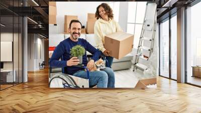 Middle age hispanic couple smiling happy. Man sitting on wheelchair with dog on his legs and woman holding cardboard box at new home. Wall mural