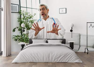 Middle age hispanic man wearing doctor uniform and stethoscope at waiting room afraid and terrified with fear expression stop gesture with hands, shouting in shock. panic concept. Wall mural