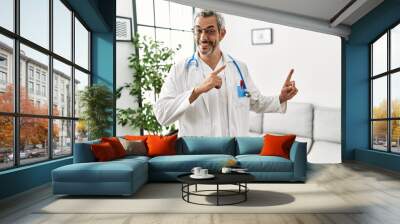 Middle age hispanic man wearing doctor uniform and stethoscope at waiting room smiling and looking at the camera pointing with two hands and fingers to the side. Wall mural