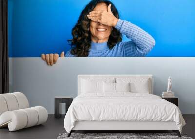 Middle age hispanic woman holding blank empty banner smiling and laughing with hand on face covering eyes for surprise. blind concept. Wall mural