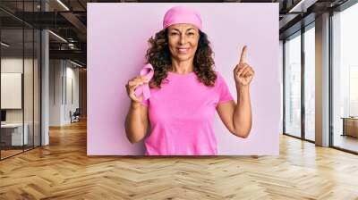 Middle age hispanic woman holding pink cancer ribbon smiling happy pointing with hand and finger to the side Wall mural