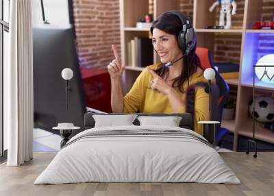 Middle age hispanic woman playing video games using headphones smiling and looking at the camera pointing with two hands and fingers to the side. Wall mural