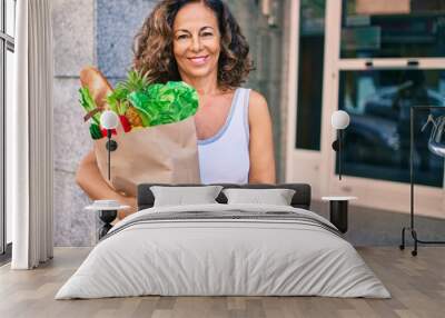 Middle age hispanic woman smiling happy holding a grocery shopping bag full of groceries at the city. Wall mural