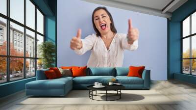 Middle age hispanic woman standing over blue background approving doing positive gesture with hand, thumbs up smiling and happy for success. winner gesture. Wall mural