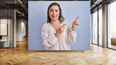 Middle age hispanic woman standing over blue background smiling and looking at the camera pointing with two hands and fingers to the side. Wall mural