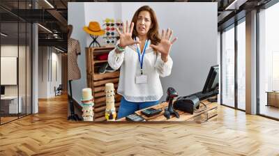 Middle age hispanic woman working as manager at retail boutique afraid and terrified with fear expression stop gesture with hands, shouting in shock. panic concept. Wall mural