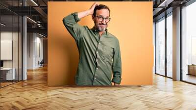 Middle age hoary man wearing casual green shirt and glasses over isolated yellow background confuse and wonder about question. Uncertain with doubt, thinking with hand on head. Pensive concept. Wall mural