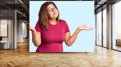 Middle age latin woman wearing casual clothes and glasses clueless and confused expression with arms and hands raised. doubt concept. Wall mural