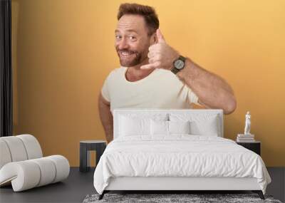 Middle age man with beard standing over yellow background smiling doing phone gesture with hand and fingers like talking on the telephone. communicating concepts. Wall mural