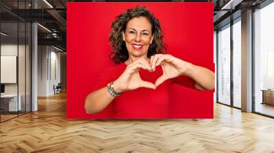 Middle age senior brunette woman wearing casual t-shirt standing over red background smiling in love doing heart symbol shape with hands. Romantic concept. Wall mural