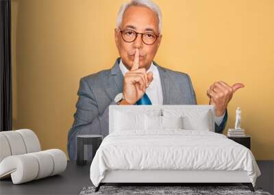 Middle age senior grey-haired handsome business man wearing glasses over yellow background asking to be quiet with finger on lips pointing with hand to the side. Silence and secret concept. Wall mural