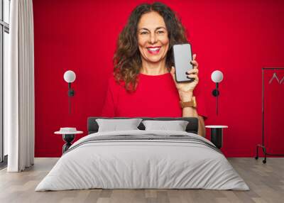 Middle age senior woman showing screen of smartphone over red isolated background with a happy face standing and smiling with a confident smile showing teeth Wall mural