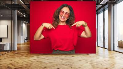 Middle age senior woman wearing cute heart shaped glasses over red isolated background looking confident with smile on face, pointing oneself with fingers proud and happy. Wall mural