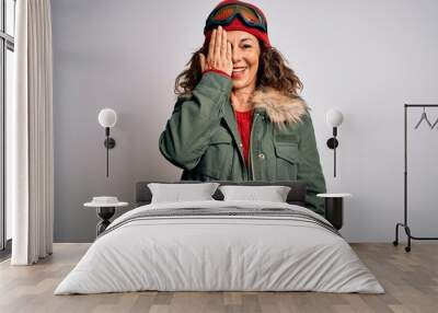 Middle age skier woman wearing snow sportswear and ski goggles over white background covering one eye with hand, confident smile on face and surprise emotion. Wall mural