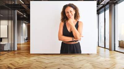 Middle age woman wearing black casual dress standing over isolated white background looking confident at the camera smiling with crossed arms and hand raised on chin. Thinking positive. Wall mural