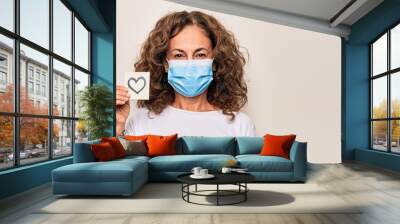 Middle age woman wearing coronavirus protection mask and holding love reminder on paper note looking positive and happy standing and smiling with a confident smile showing teeth Wall mural