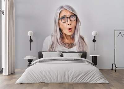 Middle age woman with grey hair standing over white background in shock face, looking skeptical and sarcastic, surprised with open mouth Wall mural