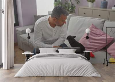 Middle-aged hispanic man sitting in a cozy living room, attentively interacting with his chihuahua dog on a gray sofa with pink pillows. Wall mural