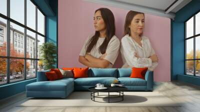 Mother and daughter standing back to back with arms crossed against a pink background, showcasing family tension and strong emotions between two women Wall mural