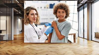 Mother and son wearing doctor uniform putting band aid on child arm at clinic Wall mural