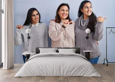 Mother and two daughters standing over blue background smiling with happy face looking and pointing to the side with thumb up. Wall mural