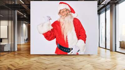 Old senior man with grey hair and long beard wearing santa claus costume with suspenders smiling with happy face looking and pointing to the side with thumb up. Wall mural