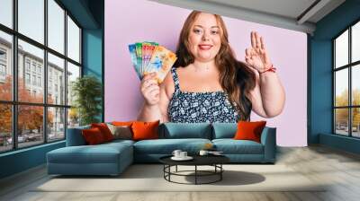 Plus size caucasian young woman holding swiss franc banknotes doing ok sign with fingers, smiling friendly gesturing excellent symbol Wall mural