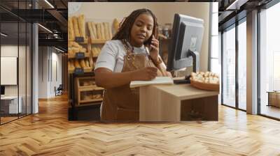 Plus size woman working in a bakery shop, taking notes, and talking on the phone while standing near a computer with a basket of pastries in the background Wall mural