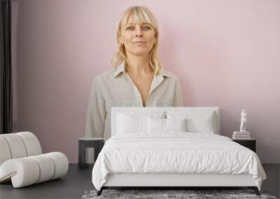 Portrait of a confident middle-aged woman with blonde hair, dressed casually, standing against a pink background. Wall mural