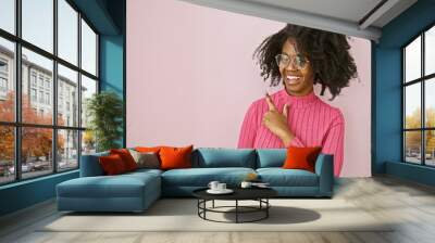 Portrait of a smiling african american woman in glasses on a pink background pointing to empty space. Wall mural