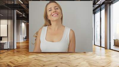 Portrait of a smiling blonde woman in a white sleeveless top against a grey background Wall mural