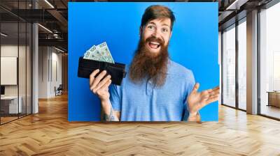 Redhead man with long beard holding wallet with polish zloty banknotes celebrating achievement with happy smile and winner expression with raised hand Wall mural