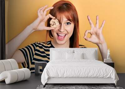 Redhead young woman holding cookie close to eye doing ok sign with fingers, smiling friendly gesturing excellent symbol Wall mural