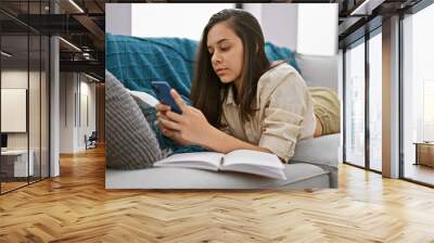 Relaxed and beautiful young hispanic woman in a deep conversation over her smartphone, lying on the living room sofa, immersed in technology at home. Wall mural