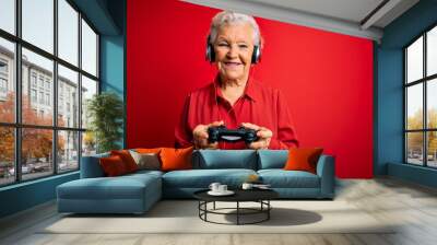 Senior beautiful grey-haired gamer woman playing video game using joystick and headphones with a happy face standing and smiling with a confident smile showing teeth Wall mural