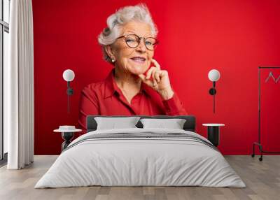 Senior beautiful grey-haired woman wearing casual shirt and glasses over red background looking confident at the camera with smile with crossed arms and hand raised on chin. Thinking positive. Wall mural