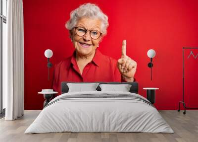 Senior beautiful grey-haired woman wearing casual shirt and glasses over red background showing and pointing up with finger number one while smiling confident and happy. Wall mural