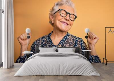 Senior beautiful grey-haired woman wearing casual shirt and glasses over yellow background very happy and excited doing winner gesture with arms raised, smiling and screaming for success. Celebration. Wall mural