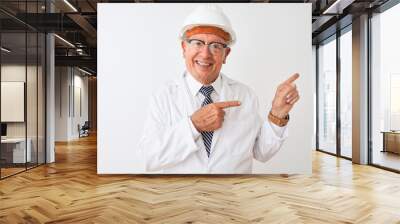 Senior grey-haired engineer man wearing coat and helmet over isolated white background smiling and looking at the camera pointing with two hands and fingers to the side. Wall mural