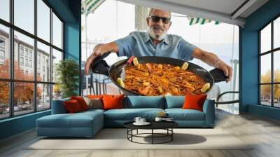 Senior grey-haired man smiling confident holding seafood paella at coffee shop terrace Wall mural