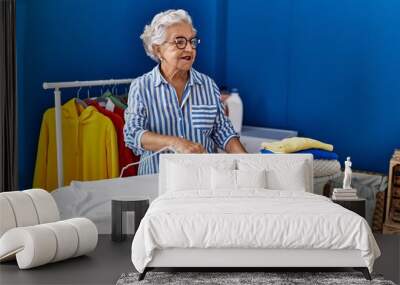 Senior grey-haired woman smiling confident ironing clothes at laundry room Wall mural