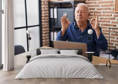 Senior man with grey hair working using computer laptop at the office relax and smiling with eyes closed doing meditation gesture with fingers. yoga concept. Wall mural